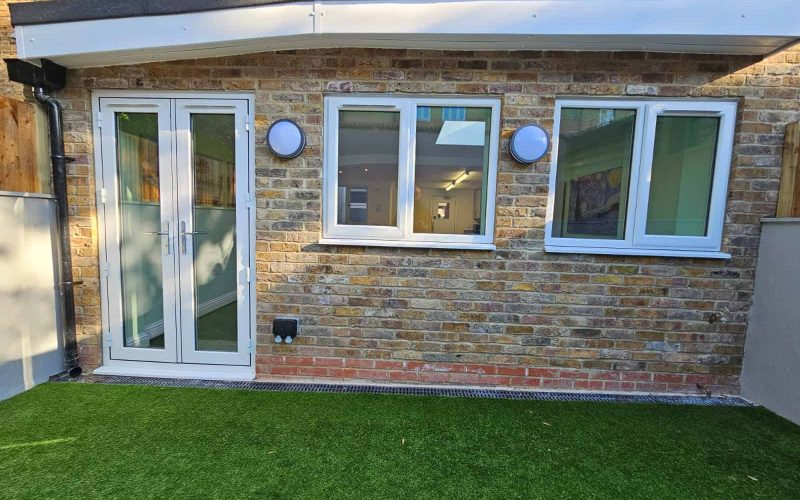 Kitchen extenson on kingsdown Road Modern brick-built garden room with white-framed French doors and windows, artificial grass lawn, and outdoor lighting.